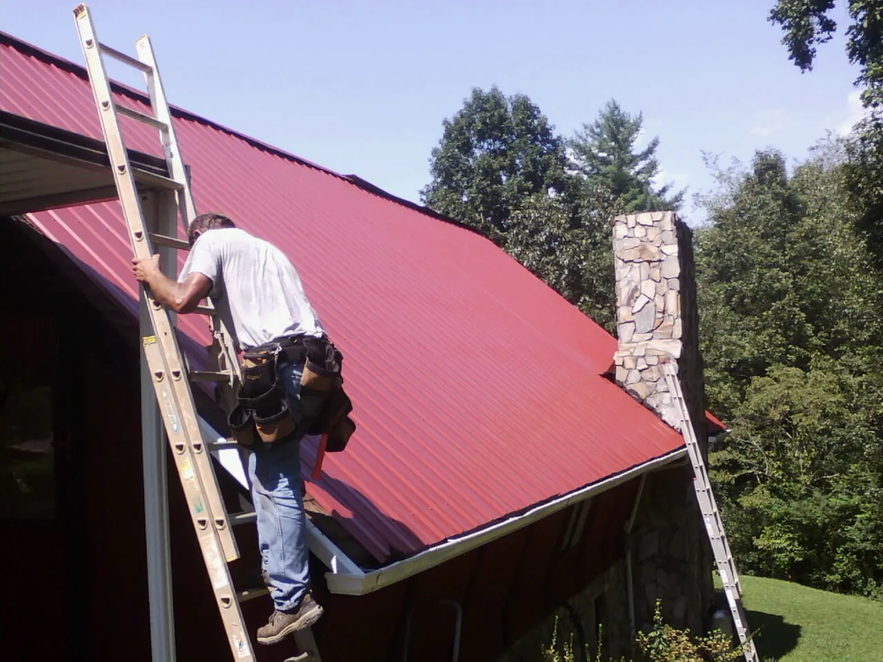 Roofer on a ladder beside a red metal roof and stone chimney — Soffit & Fascia Repair by Roofing Brothers in South Prairie